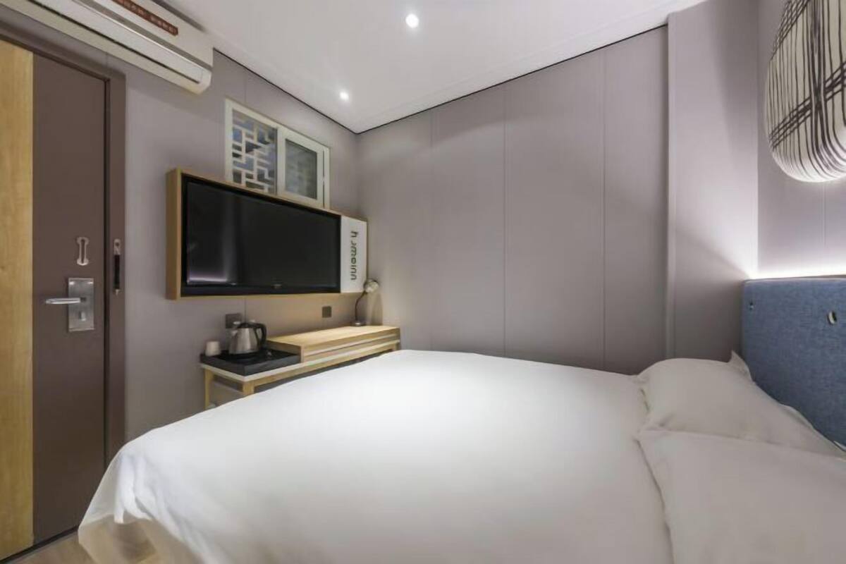 A modern hotel room with a large bed, a flat-screen TV mounted on the wall, a bedside table with a lamp, and a door with an air conditioning unit.