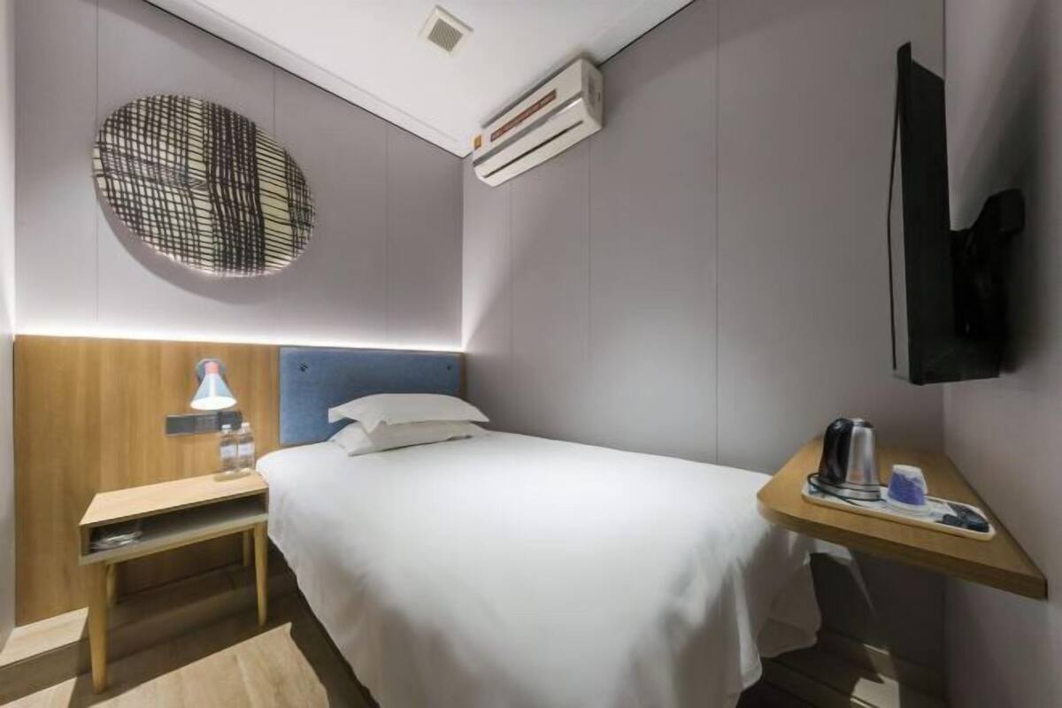 A hotel room with a bed, bedside table, lamp, air conditioning unit, TV, and a small desk with a kettle and cup.