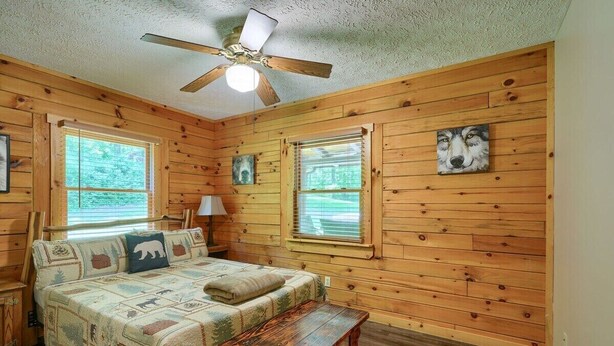 Un dormitorio estilo cabaña con cama, banco, dos ventanas, ventilador de techo y paredes de madera.