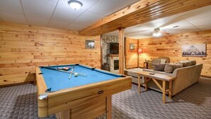 Una mesa de billar en una habitación con paredes de madera y una chimenea de piedra. Hay un sofá y una mesa con una lámpara.