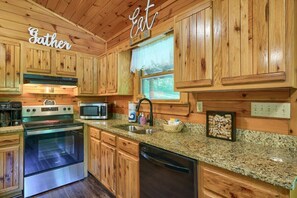 A kitchen with wooden cabinets, a granite countertop, stainless steel appliances, and a window with lace curtains. - Amazing Starr Cabin | Hocking Hills, Hot Tub, 3BR (Rockbridge)