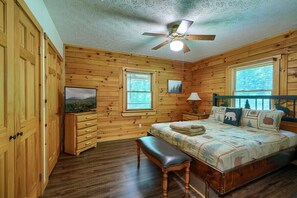 A wooden cabin bedroom with a bed, bench, ceiling fan, and two windows.