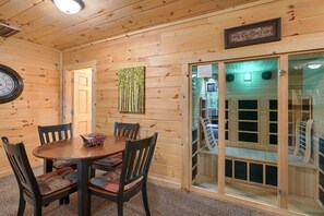 Een houten interieur met een ronde eettafel, stoelen, een klok en een sauna zichtbaar door een glazen deur.