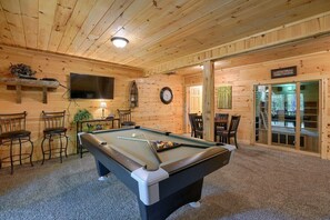 A pool table in a room with wooden walls, a clock, and a television mounted on the wall.