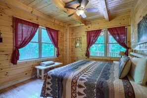 A cozy cabin room with a bed, two windows with curtains, a ceiling fan, and a bench.