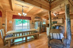 Interior de una cabaña de madera con mesa de comedor, sillas, zona de cocina y un rincón de descanso acogedor.