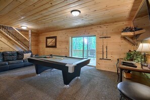 A wooden interior with a pool table, a sofa, a staircase, and a door leading outside.