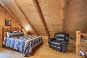 A cozy room with a wooden ceiling, a bed with patterned bedding, a leather chair, and a small table with a lamp. - Hickory House | Hocking Hills, 30 Acres, Pets (Logan)