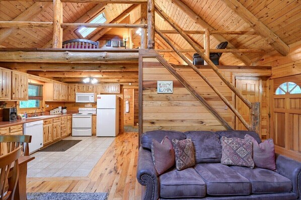 A rustic cabin interior with a kitchen, dining area, and a staircase leading to a loft.