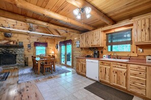 A log cabin interior with a stone fireplace, wooden dining table, and kitchen area with wooden cabinets and a dishwasher.