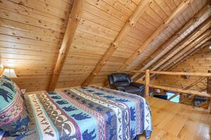 Een houten kamer met slaapgelegenheid, een schuine daklijn, beddengoed met dessin, een leren fauteuil en een zichtbare trap.