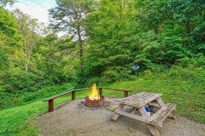 A rustic outdoor fire pit with a wooden bench and picnic table, surrounded by dense green foliage.