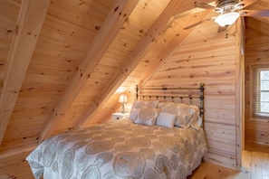 Ein Schlafzimmer mit schrägem Dach, einem Bett mit gemusterten Bettwaren, einem Nachttisch und einem Deckenventilator.