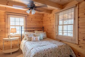 A wooden cabin bedroom with a bed, two windows with blinds, a ceiling fan, and a bedside table with a lamp.