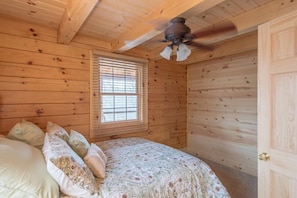 Ein Zimmer in einer Holzhütte mit einem Bett, einem Fenster mit Jalousien und einem Deckenventilator.