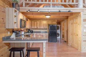 Ein Holzblockhaus-Interieur mit einer Küchenzeile, die eine Granit-Arbeitsplatte, einen schwarzen Kühlschrank und eine Waschmaschine zeigt.