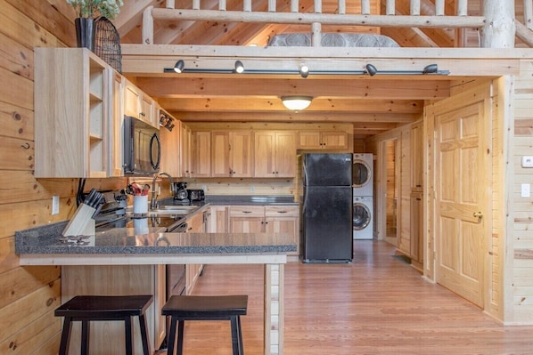 Ein Holzblockhaus-Interieur mit einer Küchenzeile, die eine Granit-Arbeitsplatte, einen schwarzen Kühlschrank und eine Waschmaschine zeigt.