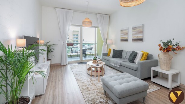 Una sala moderna con un sofá gris, una mesa de centro redonda y un reposapiés gris. Hay dos lámparas de pie, un ventanal con cortinas blancas y vistas al balcón exterior.