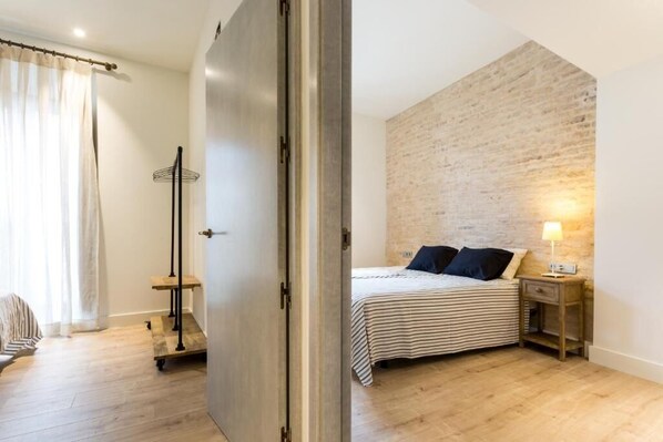 Un dormitorio con cama, mesita de noche, lámpara, suelo de madera, pared de ladrillo y una puerta.
