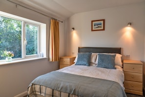 Un dormitorio con cama, mesitas de noche, ventana con cortinas, un cuadro en la pared y vistas a árboles.