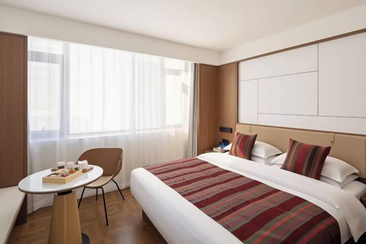 A hotel room with a bed, a small table with a tray of snacks, a chair, and a large window with curtains.