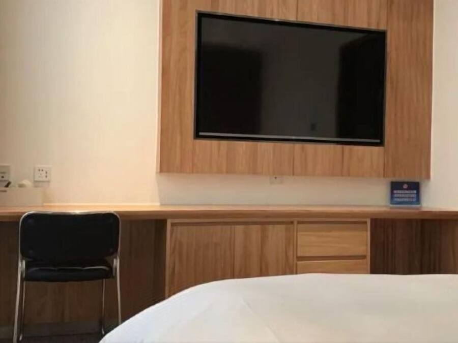 Habitación de hotel con televisión de pantalla plana montada en la pared, un escritorio con silla y una cama con colcha blanca.