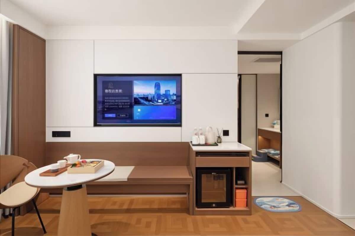A modern hotel room with a TV mounted on the wall, a small desk with a chair, a wooden bench, and a compact storage unit.
