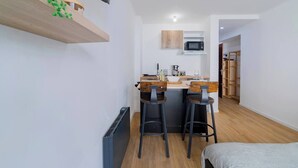 A compact kitchen with a microwave, a coffee maker, and two stools. There is a bed with a grey blanket and a wooden floor.