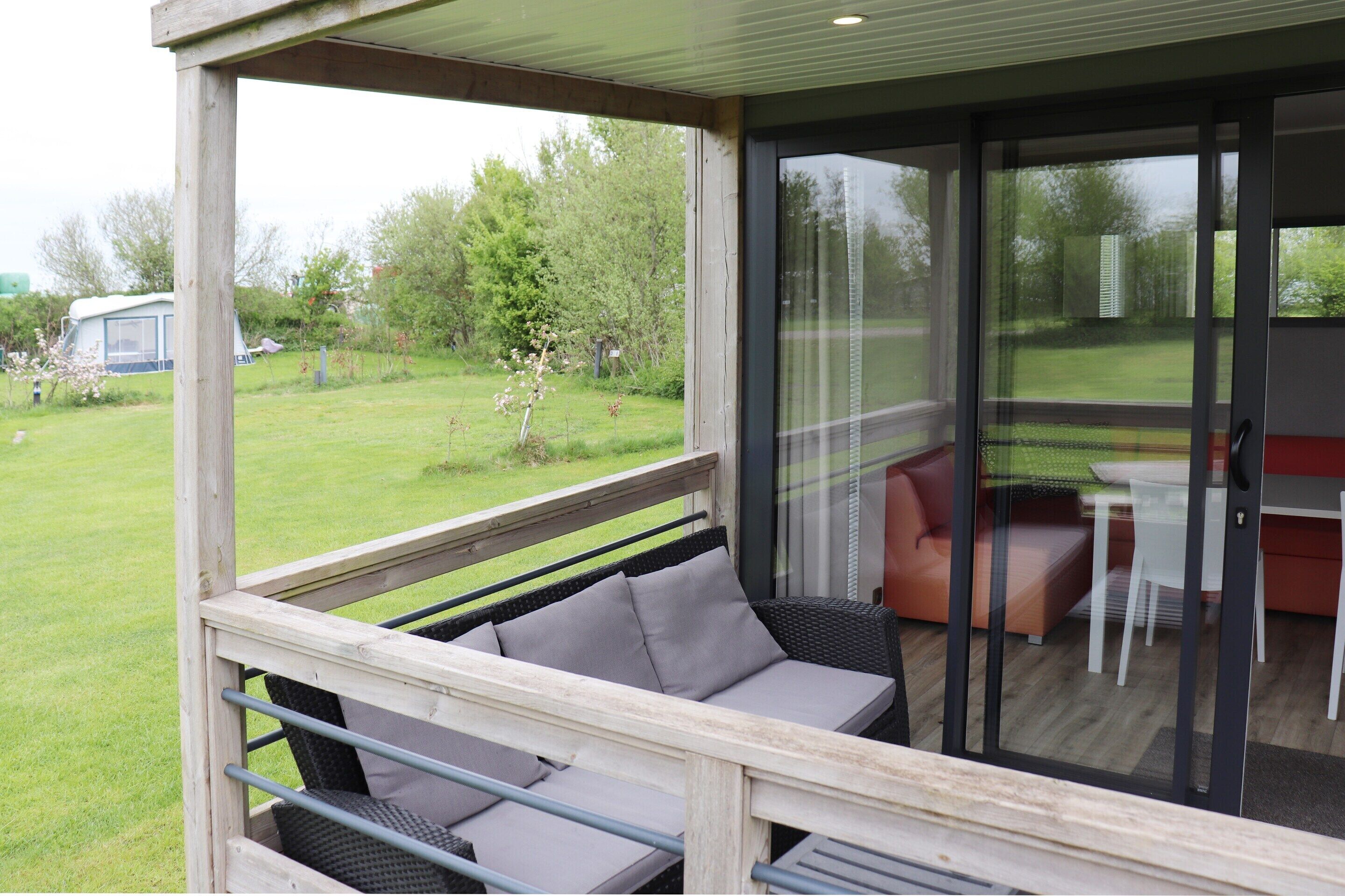 A covered outdoor seating area with a sofa and chairs, a glass door leading to an indoor space with a table and chairs, and a view of a grassy yard with trees and a parked camper.