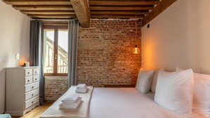Une chambre avec un mur en briques, un plafond en bois, un lit avec du linge de lit blanc, une commode et une fenêtre avec des rideaux.