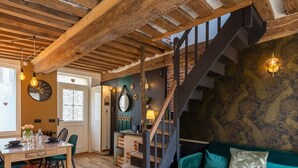 Un intérieur chaleureux avec des poutres apparentes, un escalier, un coin repas avec une table et des chaises, et un mur orné d’un motif.