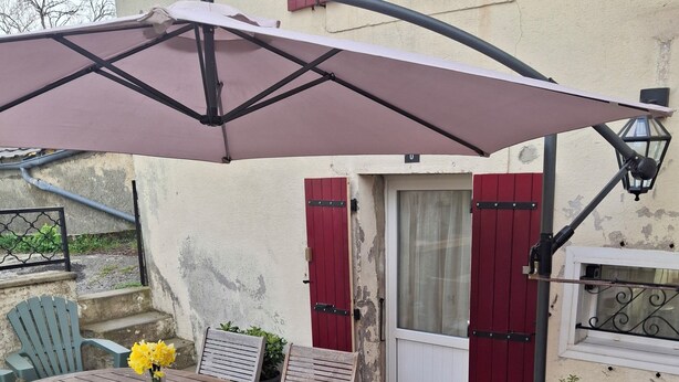 Un patio con una mesa redonda de madera, sillas y un paraguas grande. Hay una planta en maceta y una puerta con persianas rojas.