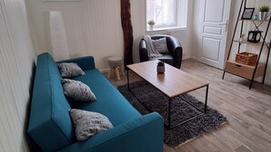 Un salón con un sofá azul, una mesa de centro de madera y una silla negra. Hay una ventana con cortinas, una puerta blanca y una planta pequeña en un estante.