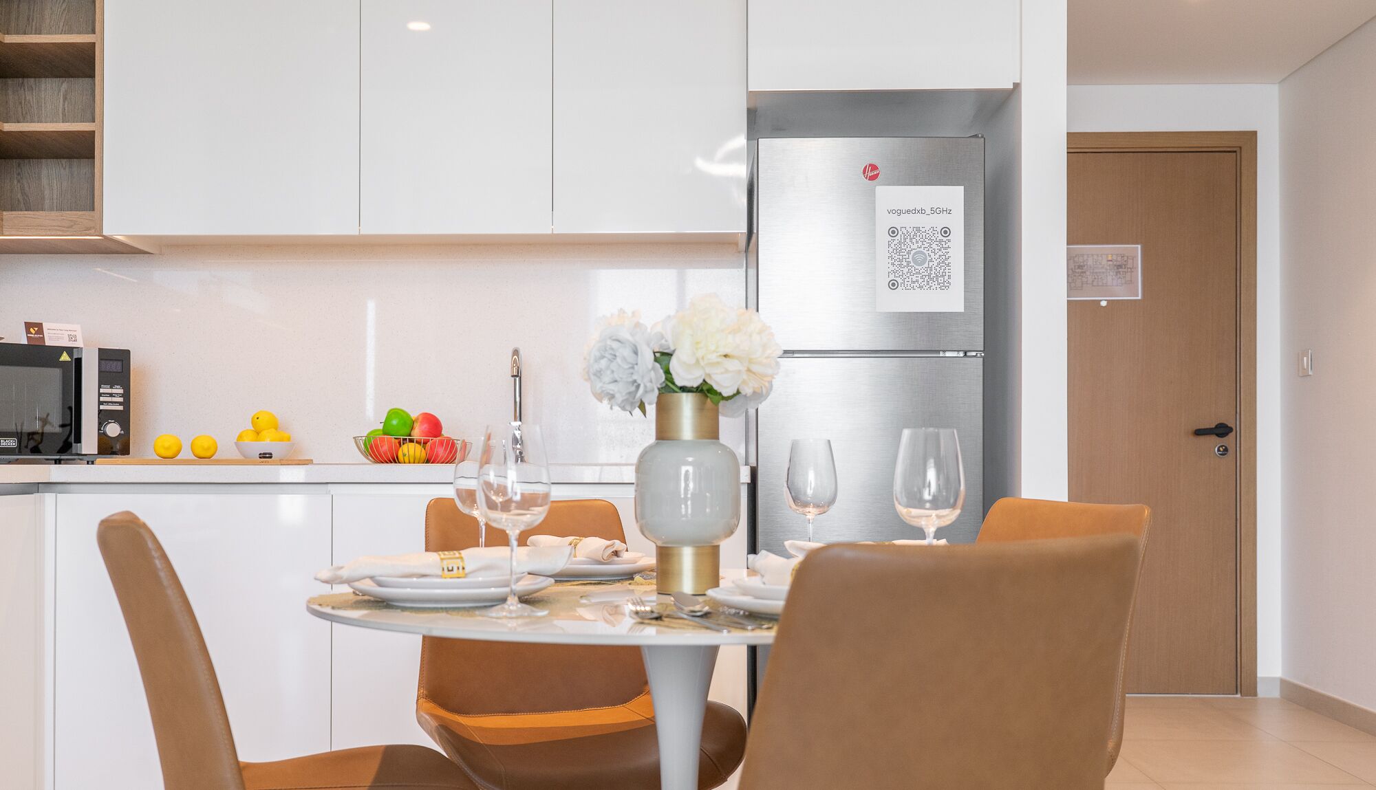Una cocina moderna con armarios blancos, un refrigerador de acero inoxidable y un comedor con una mesa redonda para dos.