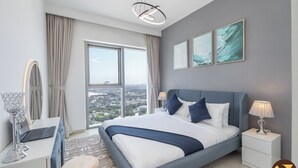 A modern bedroom with a large bed, a desk with a laptop, a vanity with a mirror, and a view of the city through large windows.
