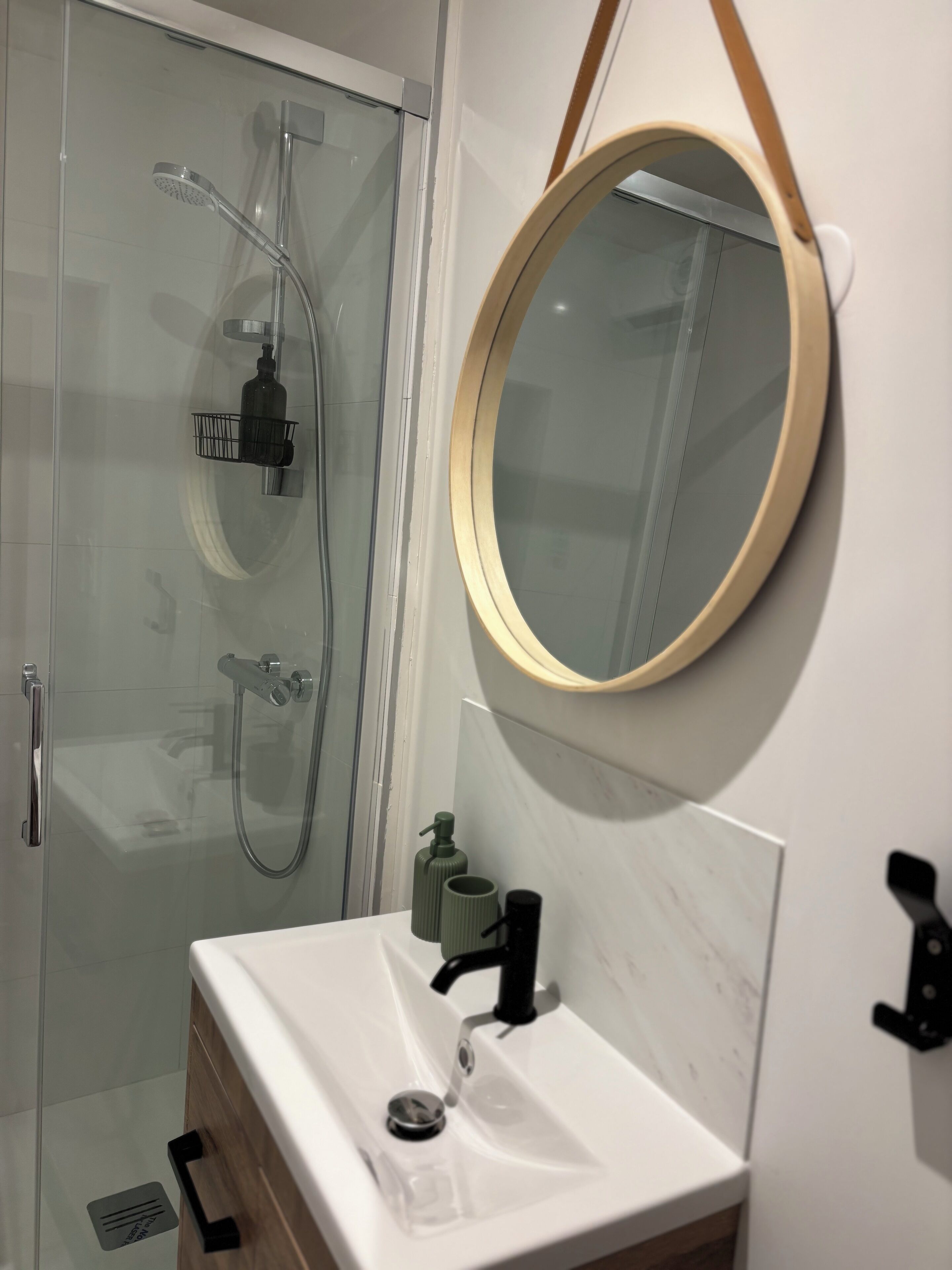 Un baño con cabina de ducha de vidrio, un lavamanos blanco con grifo negro y un espejo redondo con marco de madera.