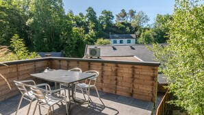 An outdoor patio with a table and chairs, surrounded by wooden walls and a view of a house in the background. - Holiday Home in La Roche Near Center (La Roche-en-Ardenne)