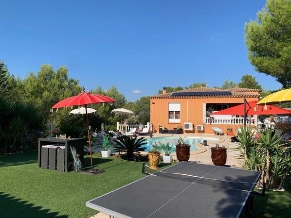 A poolside area with a ping pong table, umbrellas, and a building in the background. - Chambres d'hôtes Les Huppes (Nîmes)