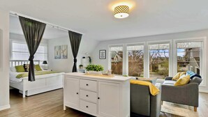 Un salon moderne avec un canapé blanc, un fauteuil gris et un meuble blanc. De grandes fenêtres offrent une vue sur l’extérieur.