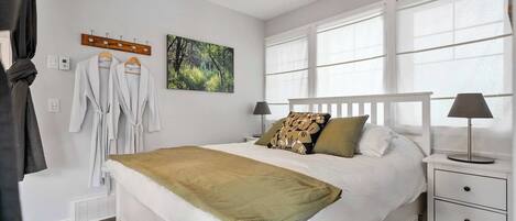 A neatly arranged bedroom with a large bed, a bedside table with a lamp, a window with curtains, and a wall-mounted coat rack with robes.