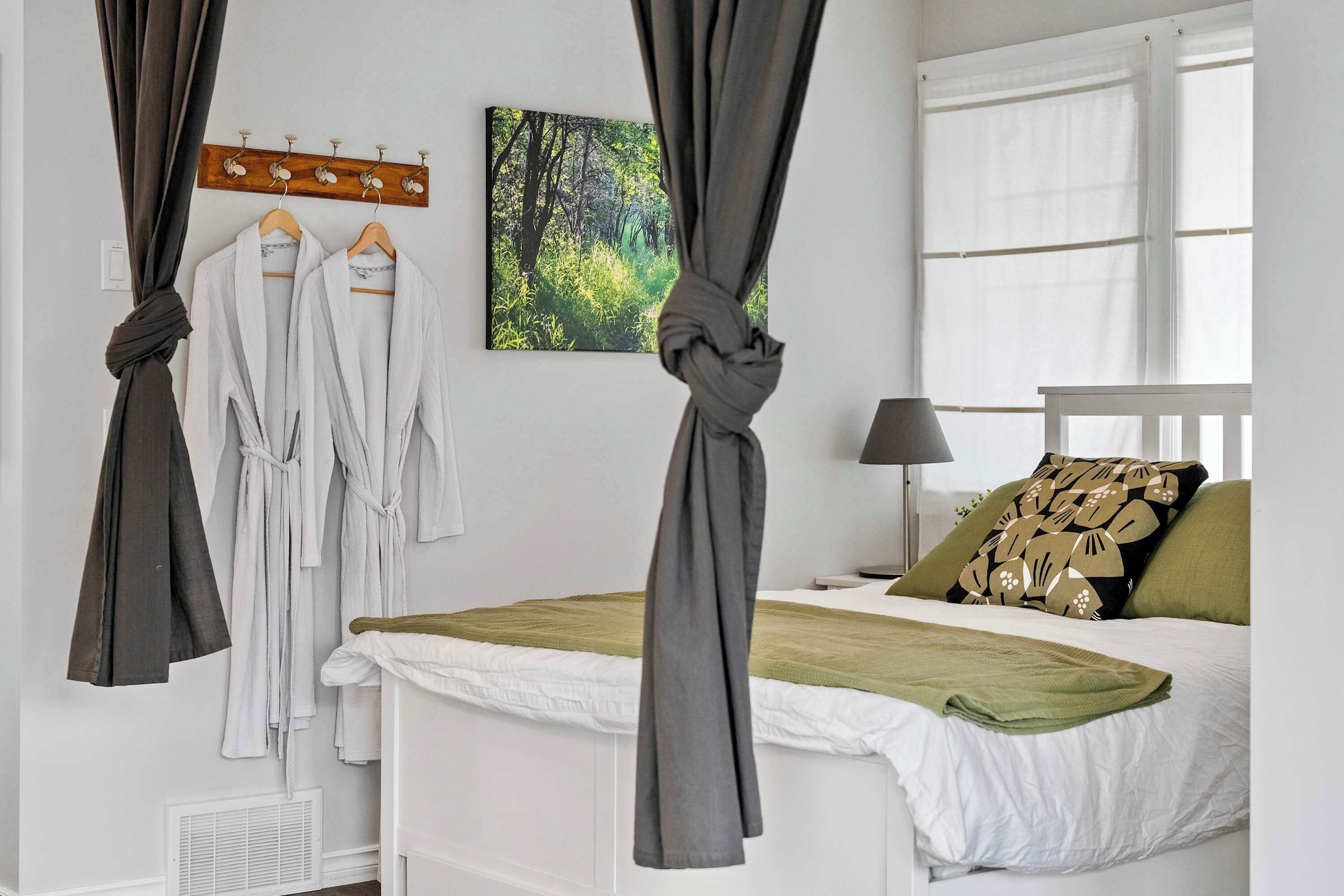 A bedroom with a bed, a window, a wall-mounted coat rack with robes, and a painting of a forest.
