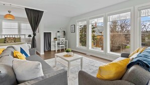 Un salon moderne avec un canapé gris, une table basse blanche et un coin repas comprenant une table et des chaises blanches. De grandes fenêtres offrent une vue sur l’extérieur.