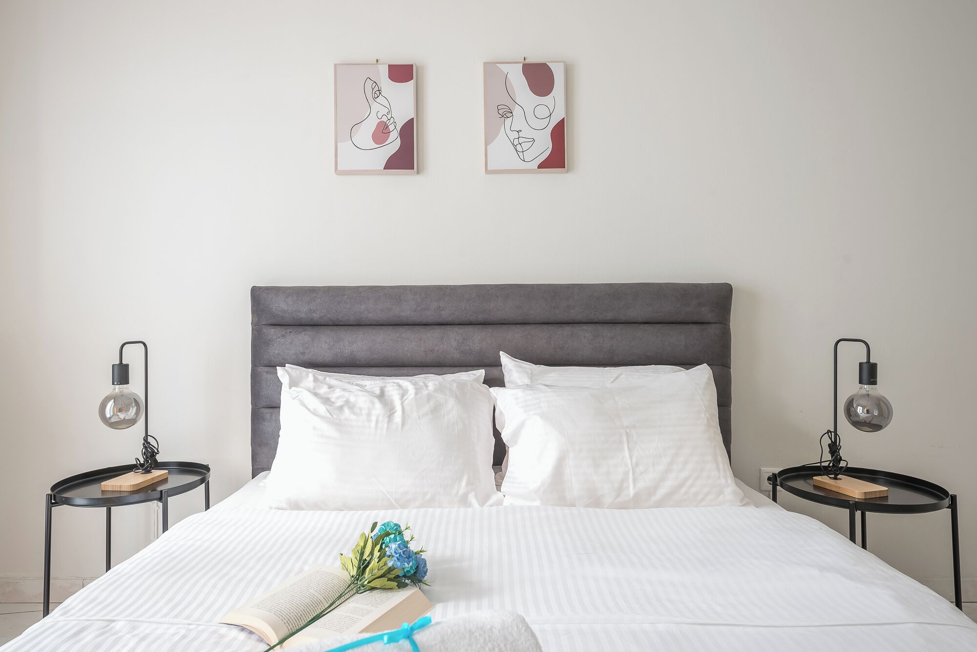 A neatly made bed with white linens, two pillows, and a book with a bouquet of flowers beside it. There are two bedside tables with lamps and a framed artwork above the bed.
