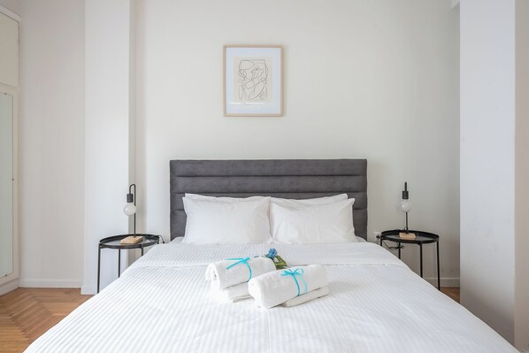 Un lit bien fait, avec du linge de lit blanc et deux oreillers, flanqué de deux tables de chevet sur lesquelles reposent des lampes, dans une chambre au style minimaliste, ornée d’une œuvre d’art encadrée au mur.