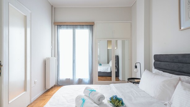 A hotel room with a bed, a bedside table, a radiator, a window with curtains, and a mirror. - 2BD - Center of Athens - 4mins Walk To Acropolis (Athina)