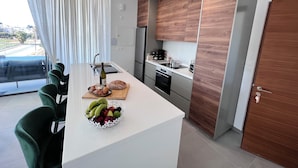 Cocina moderna con armarios de madera, una isla central con un frutero y una zona de comedor con sillas verdes.