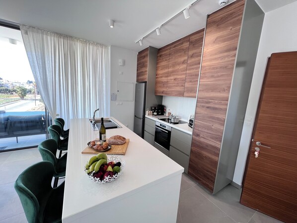 Cocina moderna con armarios de madera, una isla central con un frutero y una zona de comedor con sillas verdes.
