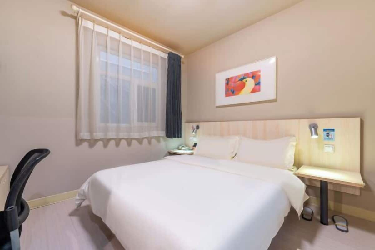 A hotel room with a bed, a desk, a chair, a window with sheer curtains, and a painting on the wall.