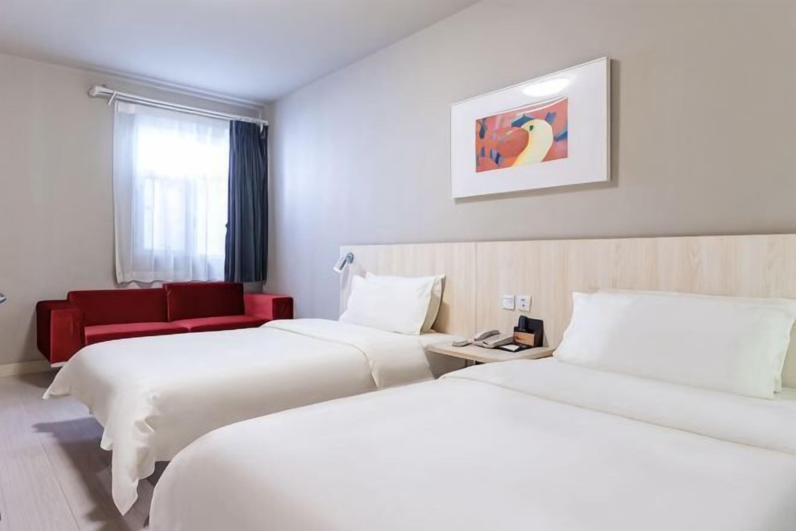 Une chambre d’hôtel avec deux lits, un canapé rouge, un tableau au mur et une fenêtre avec des rideaux.