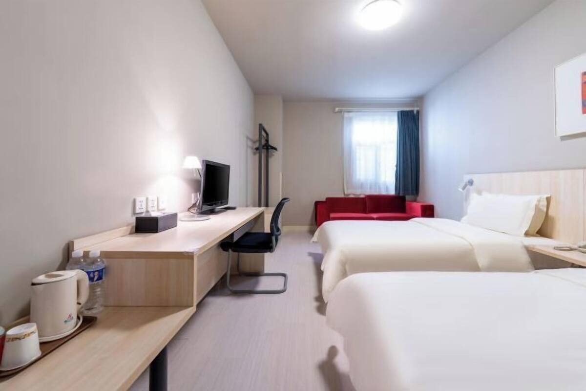A hotel room with a desk, two beds, a red chair, a television, and a window with curtains.